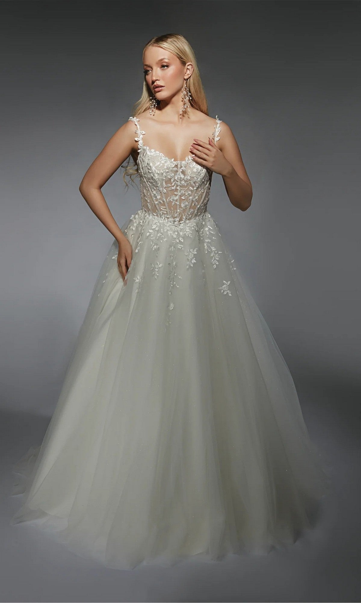 A woman in the Alyce 7130 long white dress, featuring a lace bodice and full tulle skirt, stands against a dark background, radiating effortless elegance.